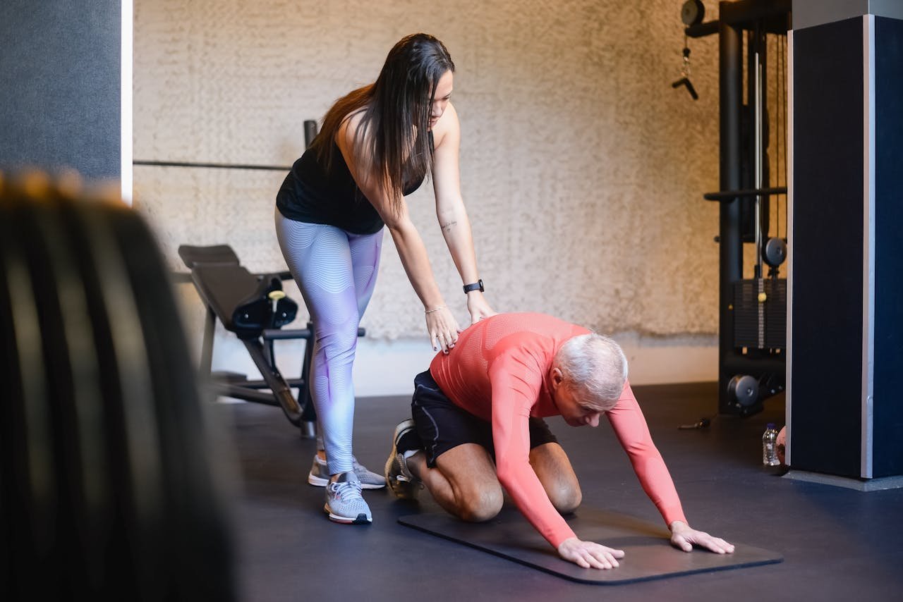 Elderly man assisted by trainer during a workout session inside a gym setting, emphasizing healthy living.